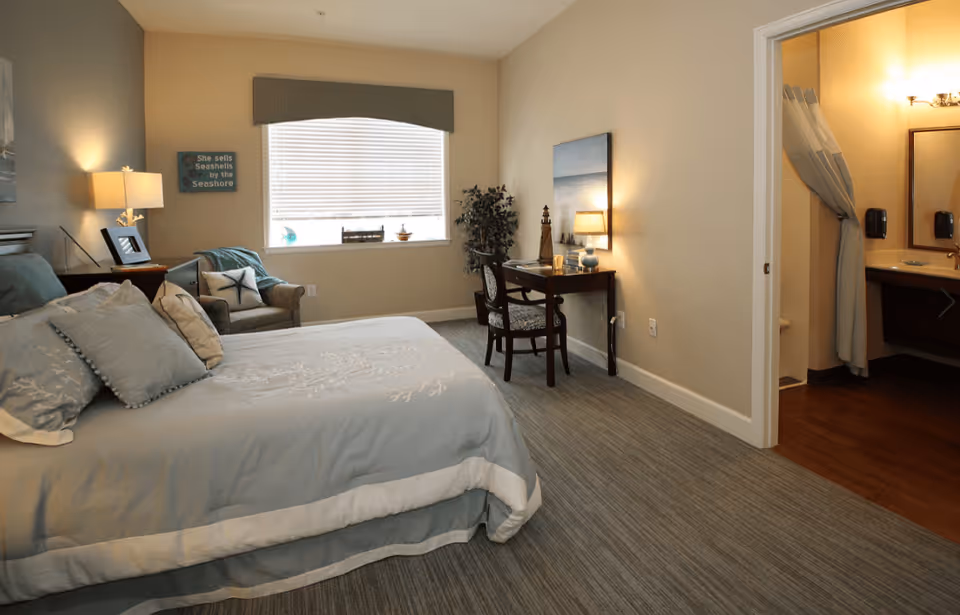 A cozy bedroom in a senior living facility with a neatly made bed featuring light blue and white bedding, a cushioned armchair near a window with blinds, a small desk with a chair and a lamp, and an open doorway leading to a bathroom with a shower curtain and sink.