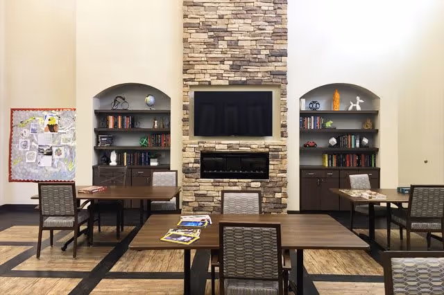 Communal dining/activity room with wooden tables and chairs facing a stone fireplace and wall-mounted TV flanked by built-in bookshelves.