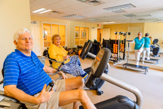 Two elderly individuals exercising on stationary bikes in a bright fitness room with mirrors on the wall. Another elderly person is seen stretching near a rack of weights and exercise equipment.