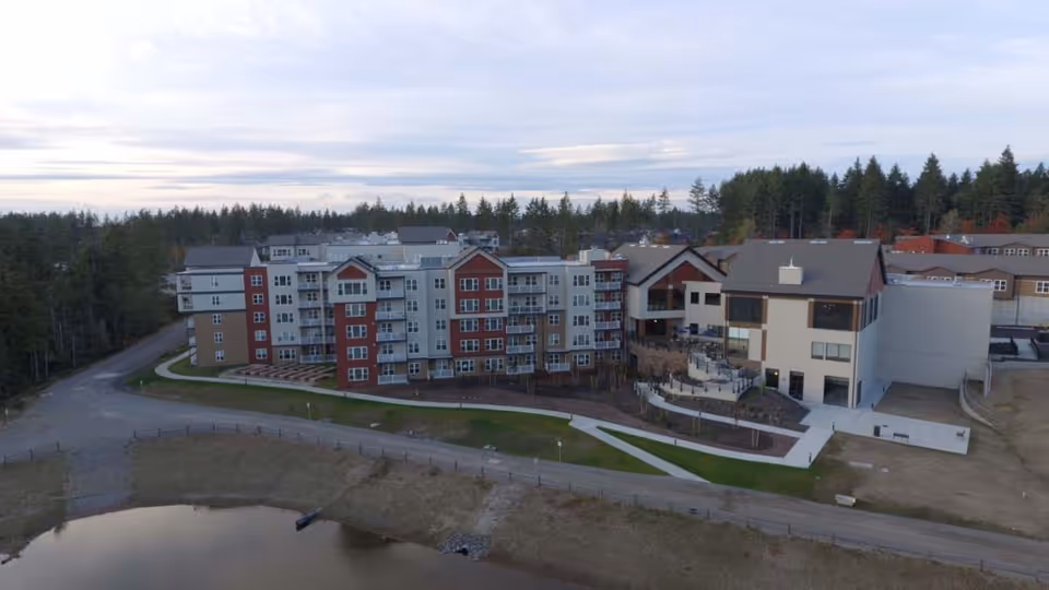 Aerial view of a large senior living facility named Heron's Key, featuring multiple connected buildings with red and beige exteriors, surrounded by trees and a small body of water in the foreground. The facility has paved walkways and landscaped areas.