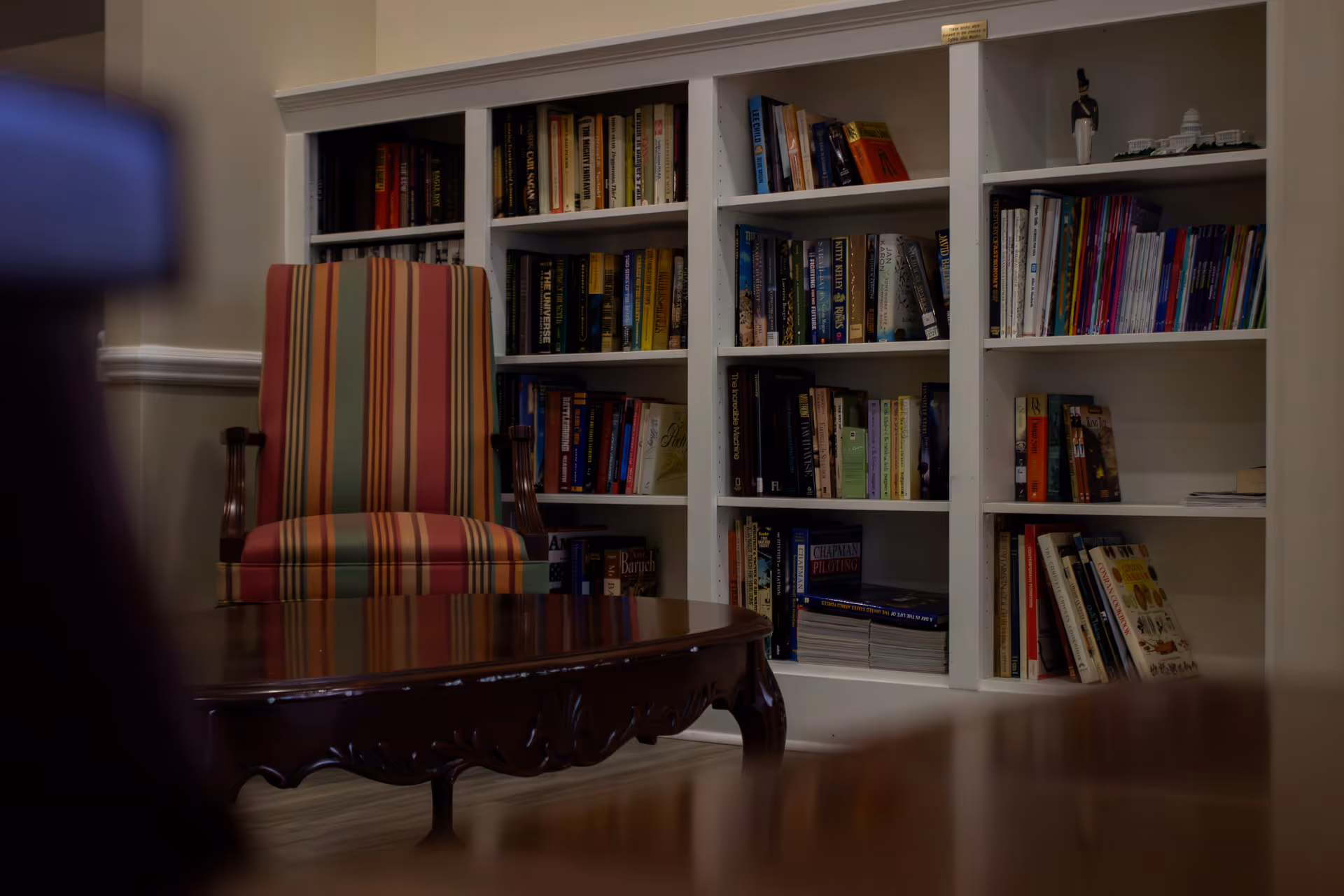 A cozy interior space featuring a wooden coffee table in the foreground, a striped upholstered armchair with red, green, and beige vertical stripes, and a white built-in bookshelf filled with various books and some decorative items.