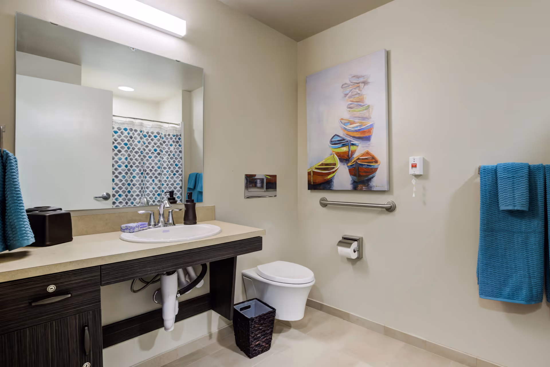 A clean and modern bathroom with a wall-mounted toilet, a sink with a countertop and dark wood cabinetry, a large mirror above the sink, a blue patterned shower curtain, blue towels hanging on a towel rack, a small wastebasket, and a colorful painting of boats on the wall.
