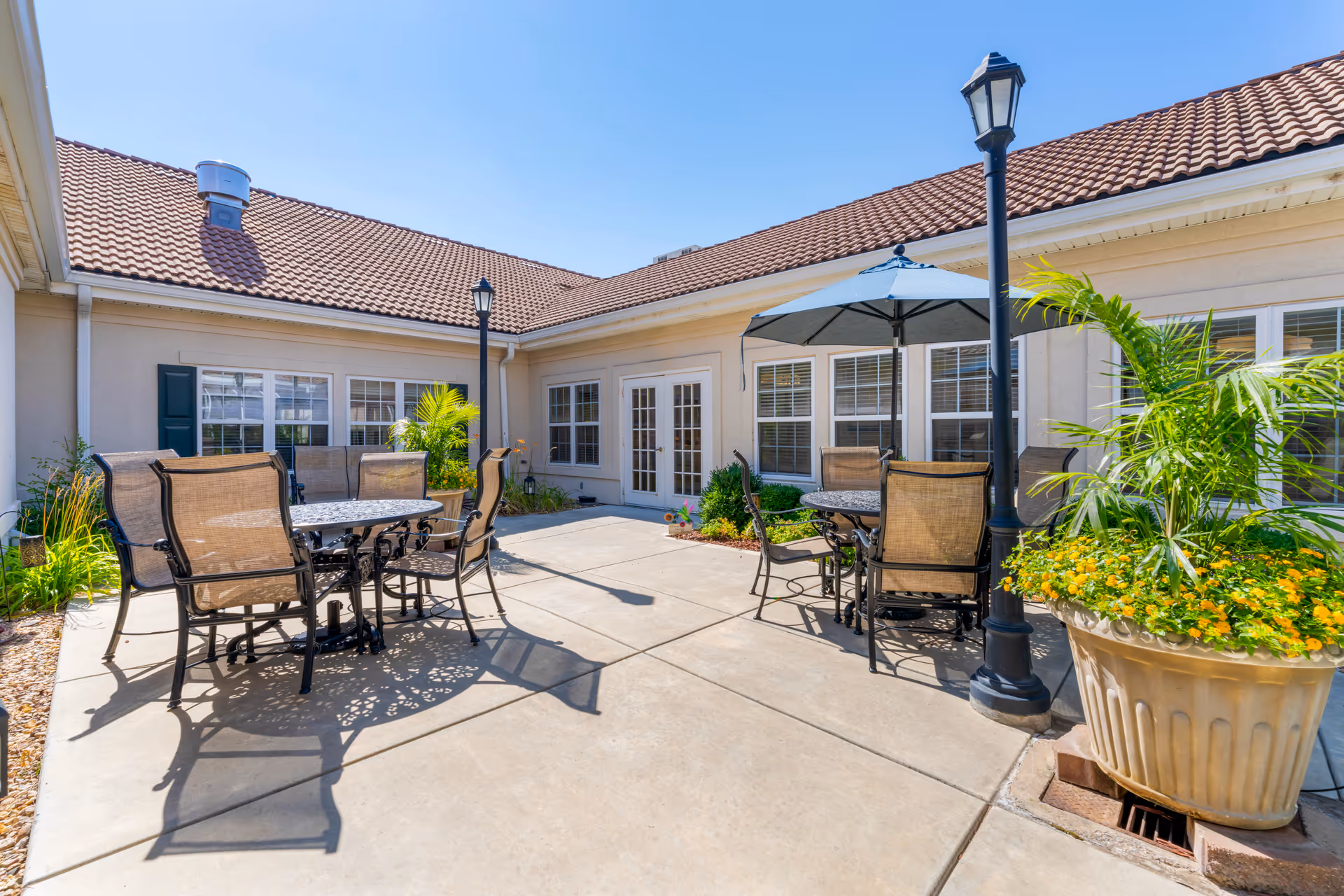 Outdoor patio area at Brookdale East Wichita with metal tables and chairs, one table shaded by a large umbrella. The patio is surrounded by a beige building with multiple windows and a tiled roof. There are potted plants and lamp posts adding to the ambiance under a clear blue sky.