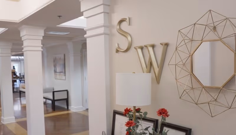 Interior view of a senior living facility with white columns and a wooden floor. On the right wall, there are large gold letters 'S' and 'W' mounted, along with a decorative geometric gold-framed mirror. Below the mirror is a table with a white lamp and a vase with red flowers. In the background, there is seating and a painting on the wall.