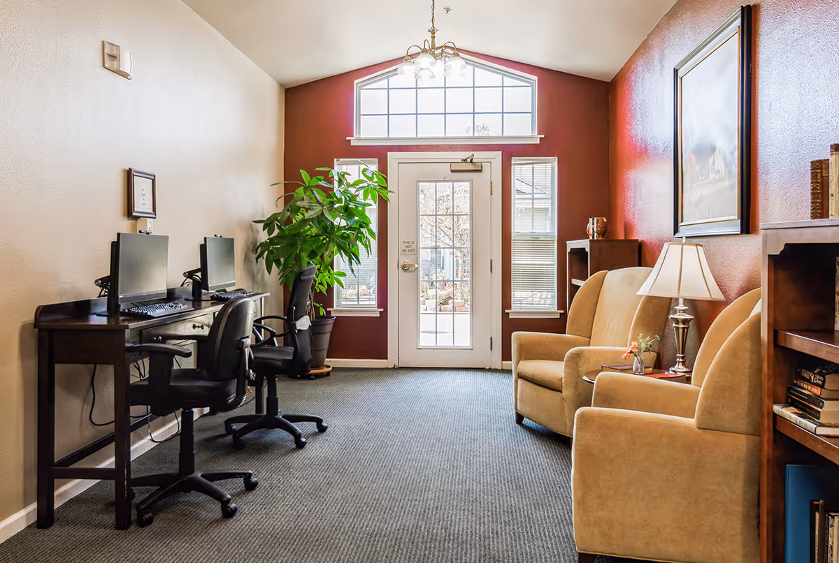 A cozy interior room with two computer desks and chairs on the left side, a large potted plant near a glass door with windows, and two beige armchairs with a small table and lamp on the right side. The walls are painted beige and red, and there is a framed picture hanging above the armchairs.