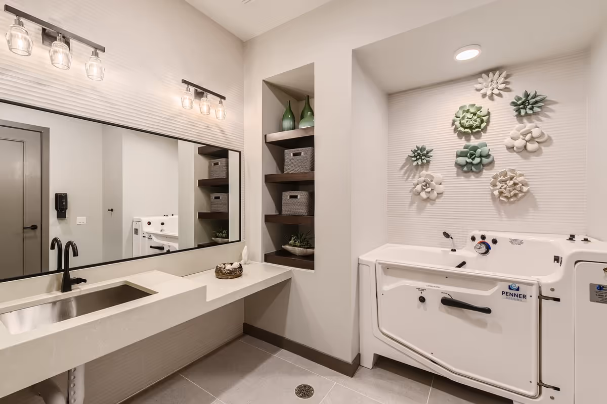 A modern bathroom with a long countertop and built-in sink, a large mirror with two sets of three-light fixtures above it, open shelving with decorative baskets and green bottles, and a white walk-in bathtub with floral wall decorations above it.