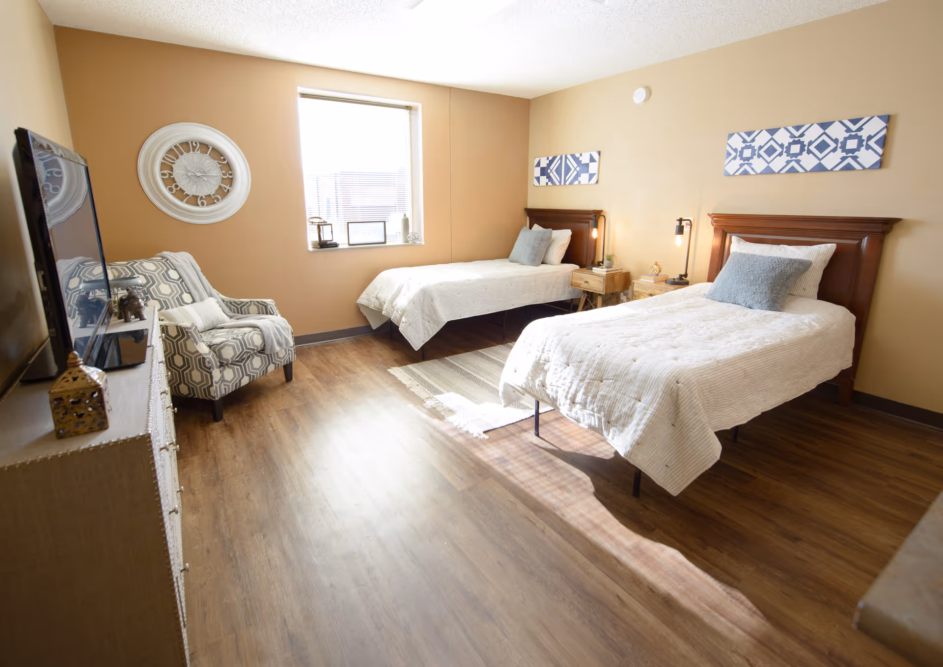 A bright senior living bedroom with two single beds each with white bedding and a gray pillow. Between the beds is a wooden nightstand with a lamp and small decor. A patterned armchair is positioned near a window with blinds, and a large wall clock hangs on the adjacent wall. The room has wood flooring and beige walls with two blue and white patterned artworks above the beds.