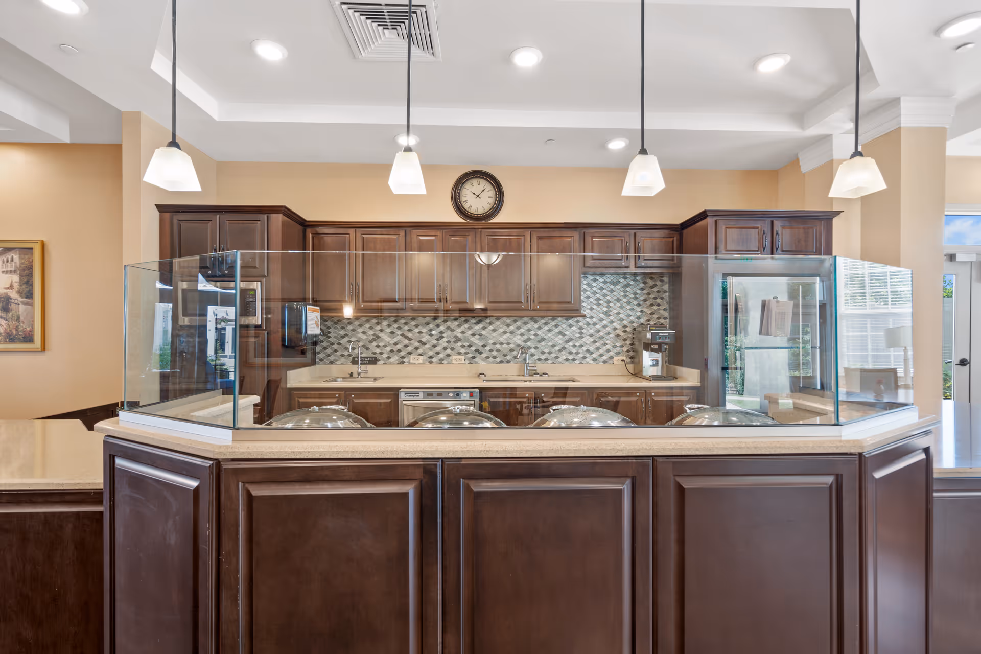 A serving counter with a glass sneeze guard in front of a kitchen featuring dark wood cabinets, pendant lights, and a wall clock.