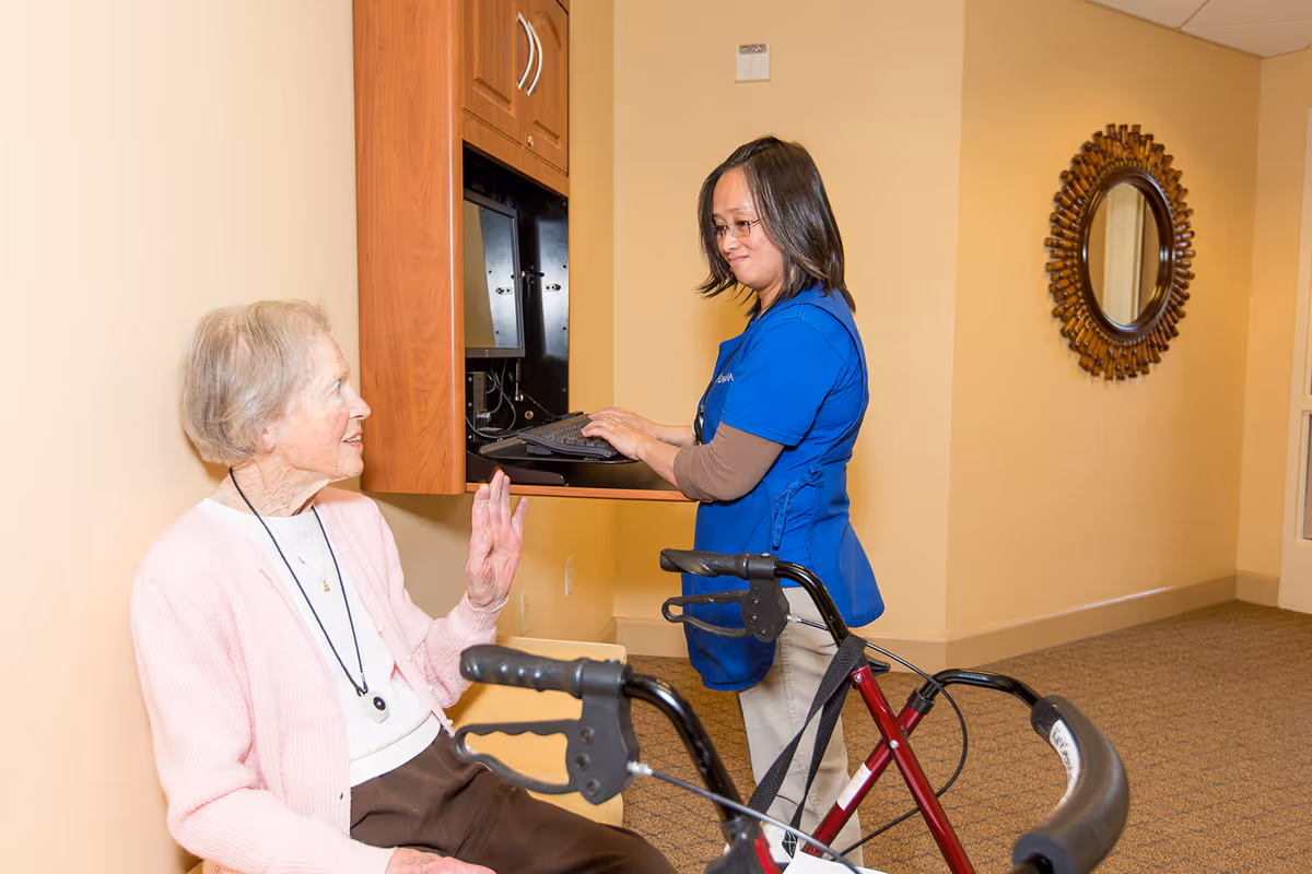 An elderly woman sitting and smiling while talking to a healthcare worker in blue scrubs who is standing and typing on a computer mounted on the wall. A red walker is positioned in front of the elderly woman. The setting appears to be a hallway or common area with beige walls and a decorative round mirror on the wall.