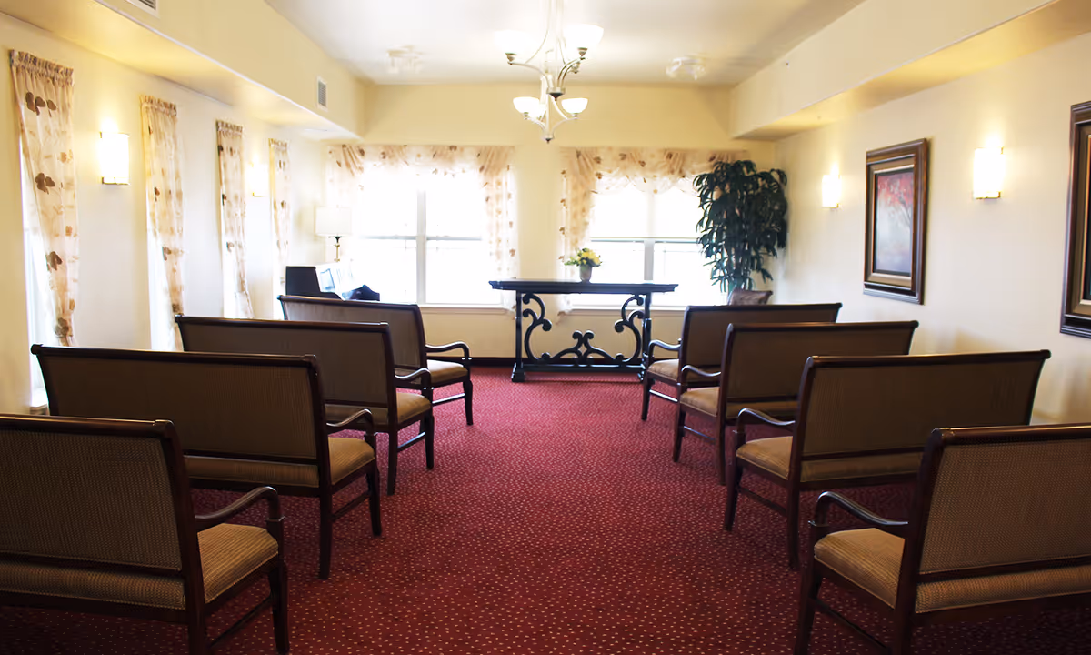 A small meeting or chapel room with rows of wooden chairs with cushioned seats arranged facing a decorative table in front of two large windows with sheer curtains. The room has beige walls, red carpet with a subtle pattern, wall sconces, framed artwork, and a large potted plant near the windows.