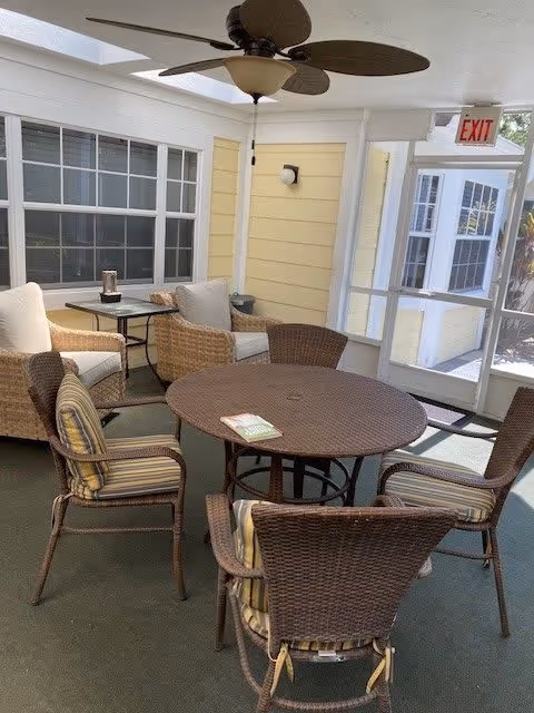 A sunroom at Discovery Commons At Bradenton featuring a round table surrounded by wicker chairs, with a ceiling fan above and large windows allowing natural light.
