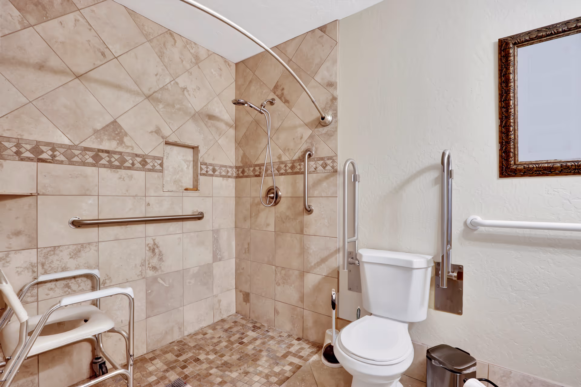 Accessible bathroom with beige tiled walls and floor, featuring a walk-in shower with a handheld showerhead, grab bars, a shower chair, a toilet with support rails, a framed mirror, and a small trash can.