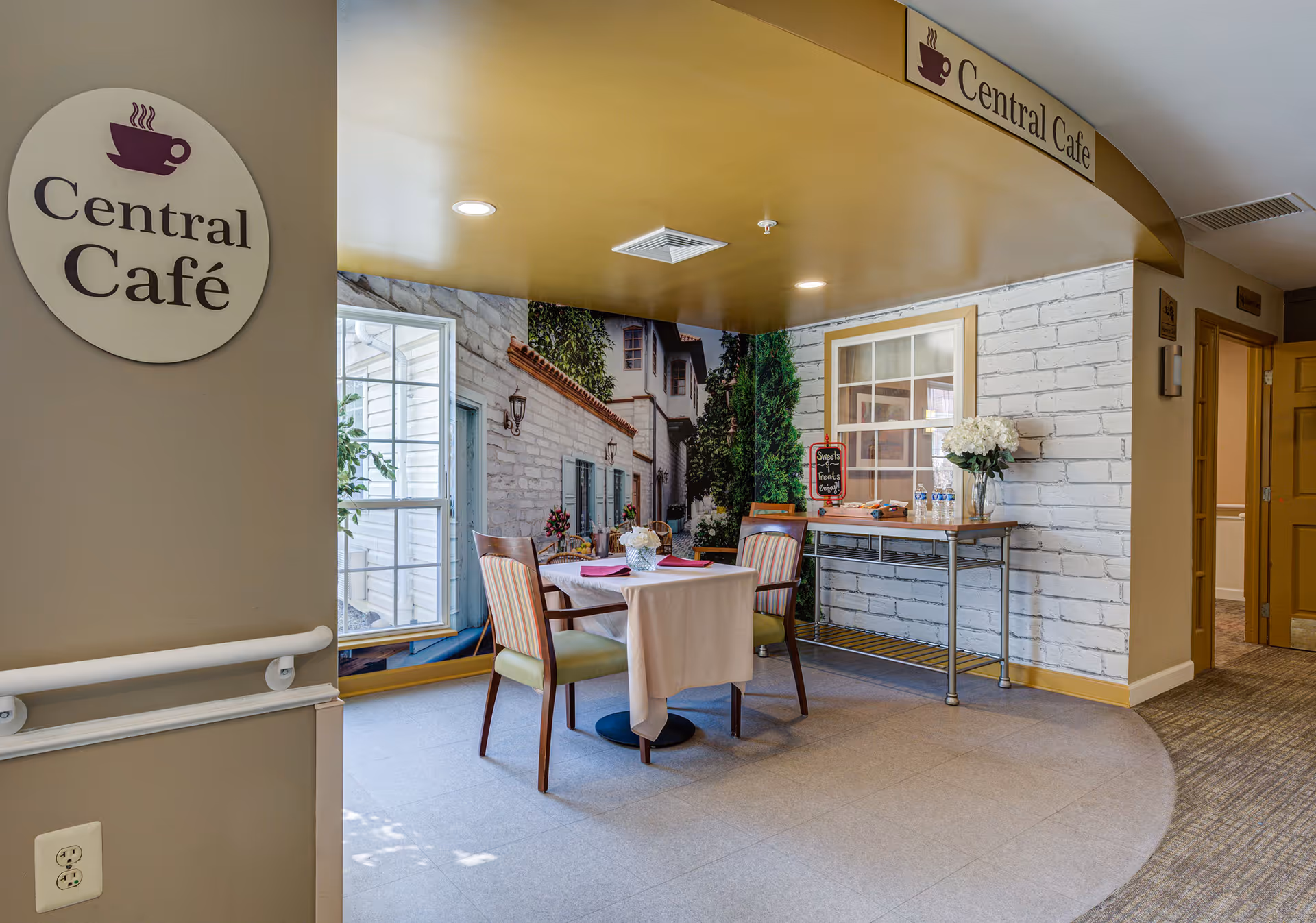 A small café area named Central Café with a round table covered with a white tablecloth and two chairs. The walls have a mural depicting a quaint outdoor scene with buildings and greenery. There is a side table with water bottles and a vase of white flowers. The ceiling is painted yellow and there are signs indicating the Central Café.