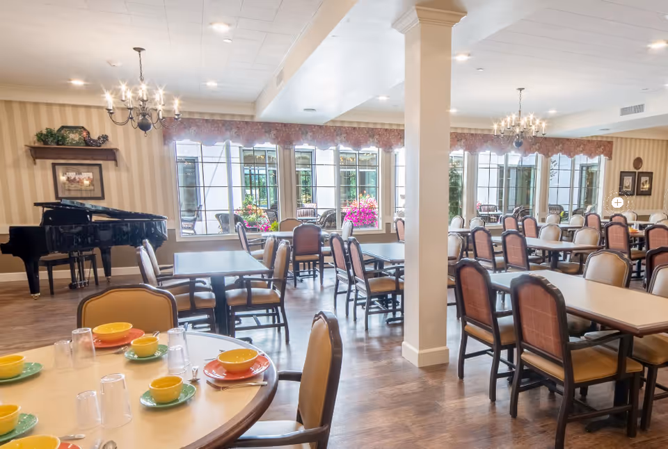 A spacious dining room with multiple tables and chairs arranged neatly. The tables are set with colorful plates, bowls, and cups. Large windows allow natural light to fill the room, and a black grand piano is positioned against one wall. The room features chandeliers hanging from the ceiling and decorative wall accents.