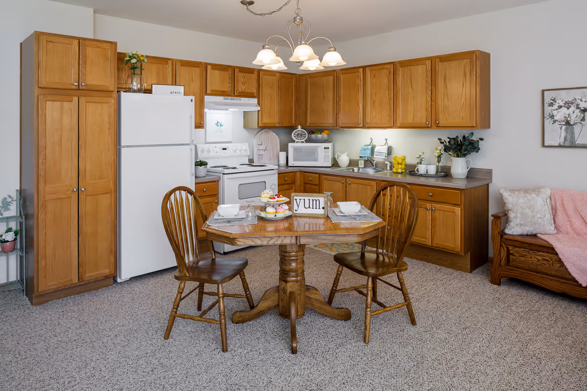 A cozy kitchen and dining area with wooden cabinets, a white refrigerator, stove, and microwave. A small wooden dining table with two chairs is set with plates, bowls, and a tiered tray of cupcakes. The table has a decorative sign that says 'yum.'. To the right, there is a wooden bench with a pink blanket and a white fluffy pillow. The room has a carpeted floor and a light fixture hanging from the ceiling.