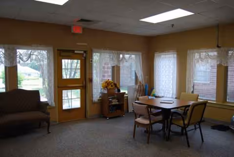 Cozy common dining area with a table, chairs, sofa, and windows in a senior facility.