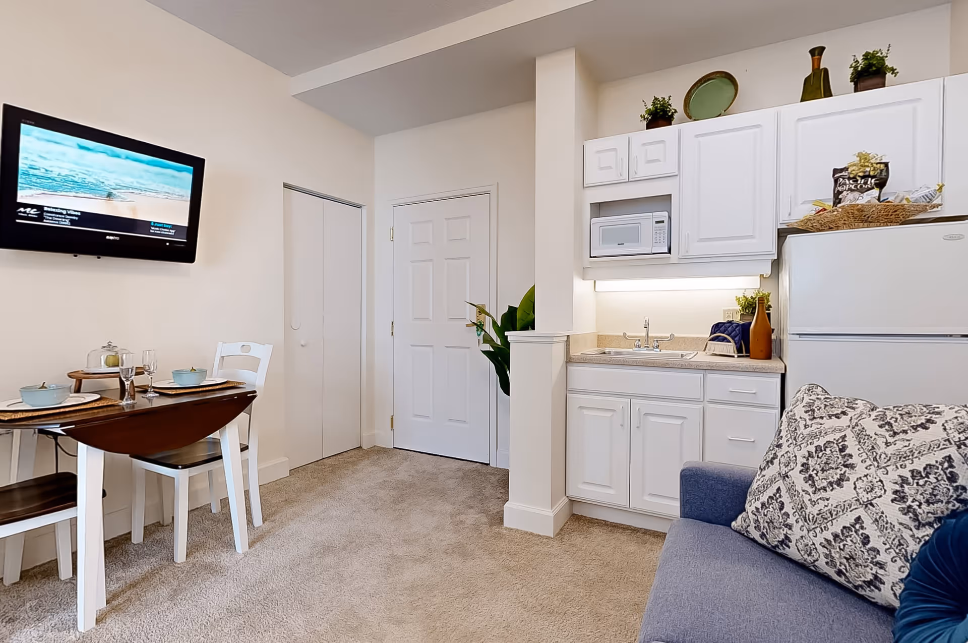 A small living space featuring a kitchenette with white cabinets, a sink, microwave, and refrigerator. To the left, there is a small dining table set for two with dishes and glasses. A flat-screen TV is mounted on the wall above the dining area. A blue couch with patterned pillows is partially visible on the right side. The room has beige carpet and neutral-colored walls.