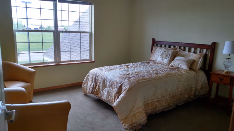 Sunlit bedroom with a double bed, nightstand and lamp, two armchairs, and a large window overlooking a grassy area.