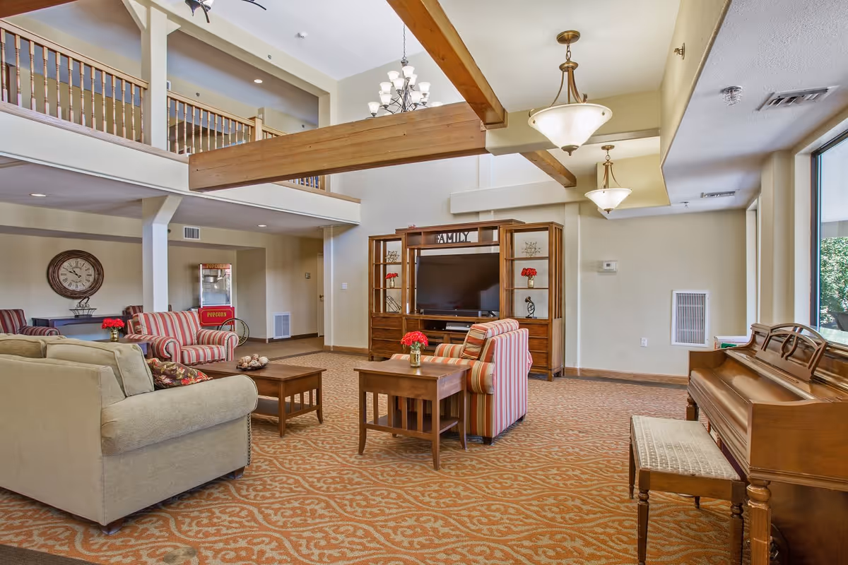 A spacious living room area in a senior living facility with beige and striped armchairs, a beige sofa, wooden coffee and side tables with red flower vases, a large wooden entertainment center with a TV, a piano with a cushioned bench, and a popcorn machine in the background. The room features high ceilings with exposed wooden beams, chandeliers, and large windows letting in natural light.