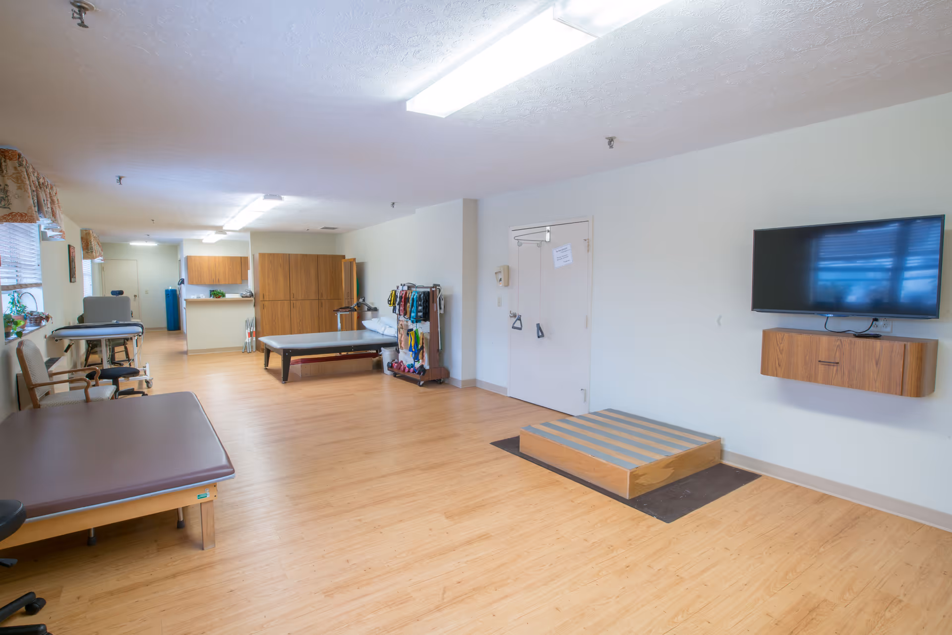 Spacious interior therapy/exercise room with treatment tables, wooden flooring, wall-mounted TV and various equipment.