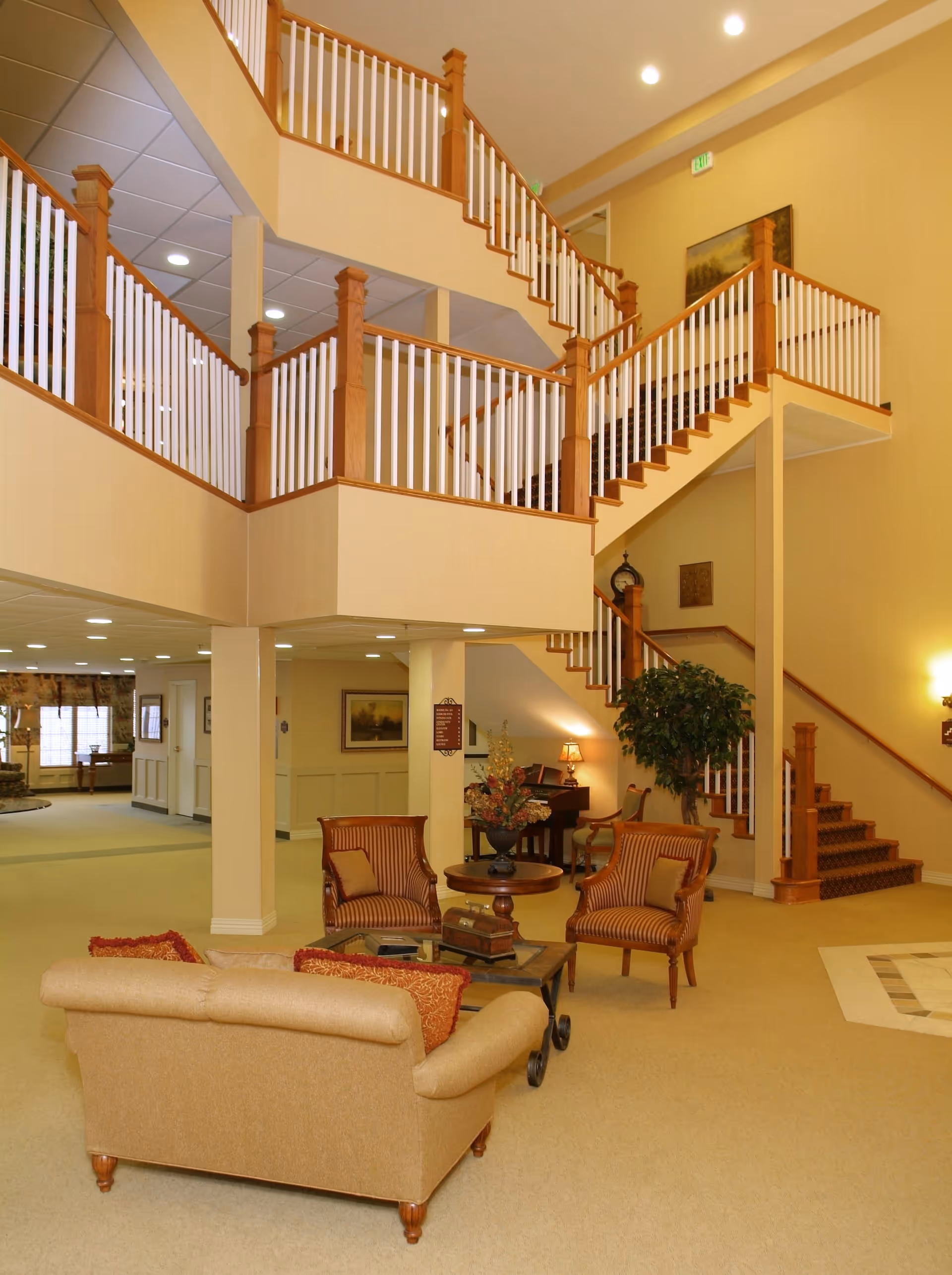 Interior view of a senior living community common area featuring a beige sofa with red cushions, two striped armchairs, a coffee table with decorative items, a staircase with wooden handrails and white balusters, a potted plant, and warm yellow walls with framed artwork and lighting fixtures.