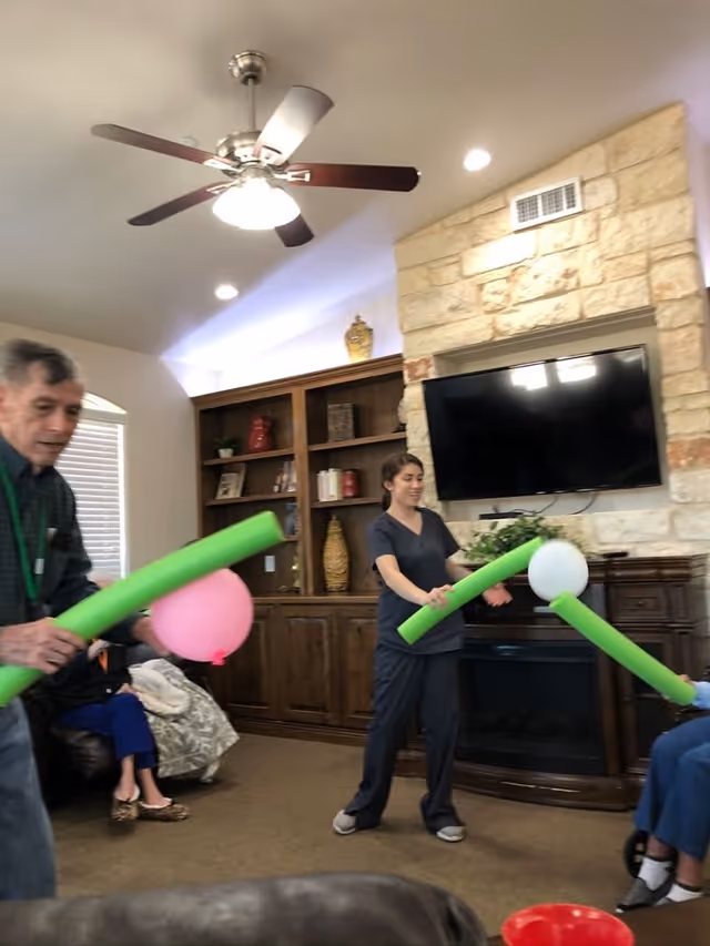 A group of people in a living room playing a game with green foam noodles and balloons. The room has a ceiling fan, a stone fireplace with a mounted TV, and wooden shelves with decorative items.
