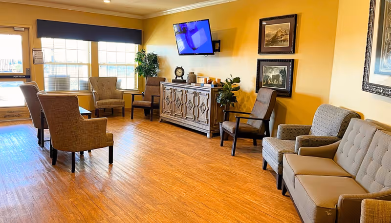 Bright senior living lounge with multiple armchairs and sofas arranged around a wooden cabinet and wall-mounted TV.