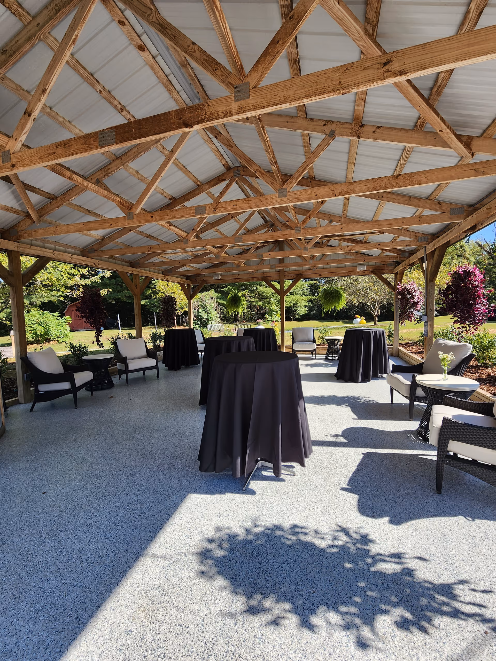 Outdoor covered patio area with wooden beams and a metal roof, featuring several high-top tables covered with black tablecloths and cushioned chairs arranged around the space. The patio overlooks a garden with green trees and shrubs under a clear blue sky.