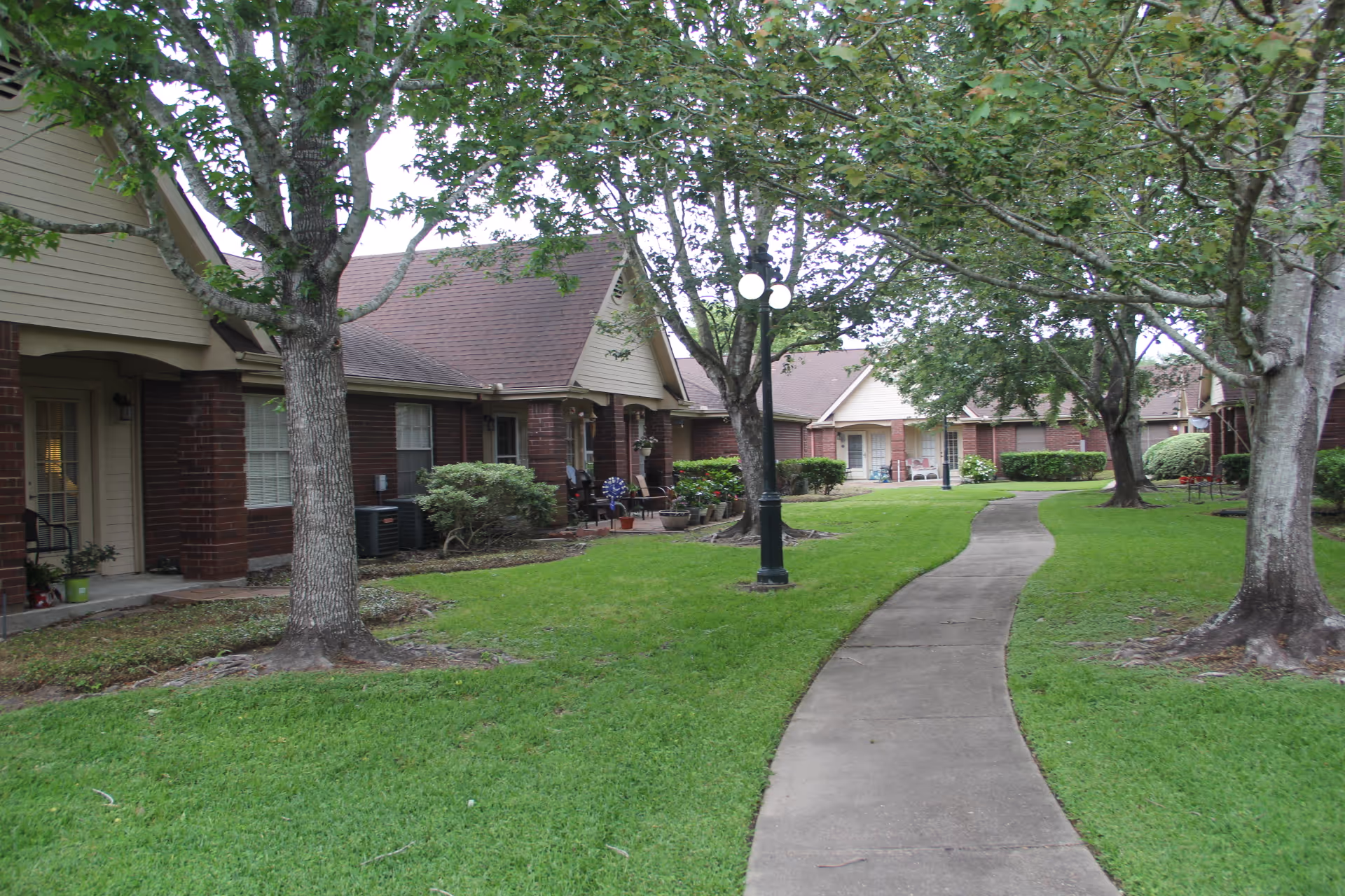 A paved walkway curves through a grassy courtyard with several mature trees and lamp posts. Surrounding the courtyard are single-story brick residential buildings with beige siding and sloped roofs. Some porches have chairs and potted plants.