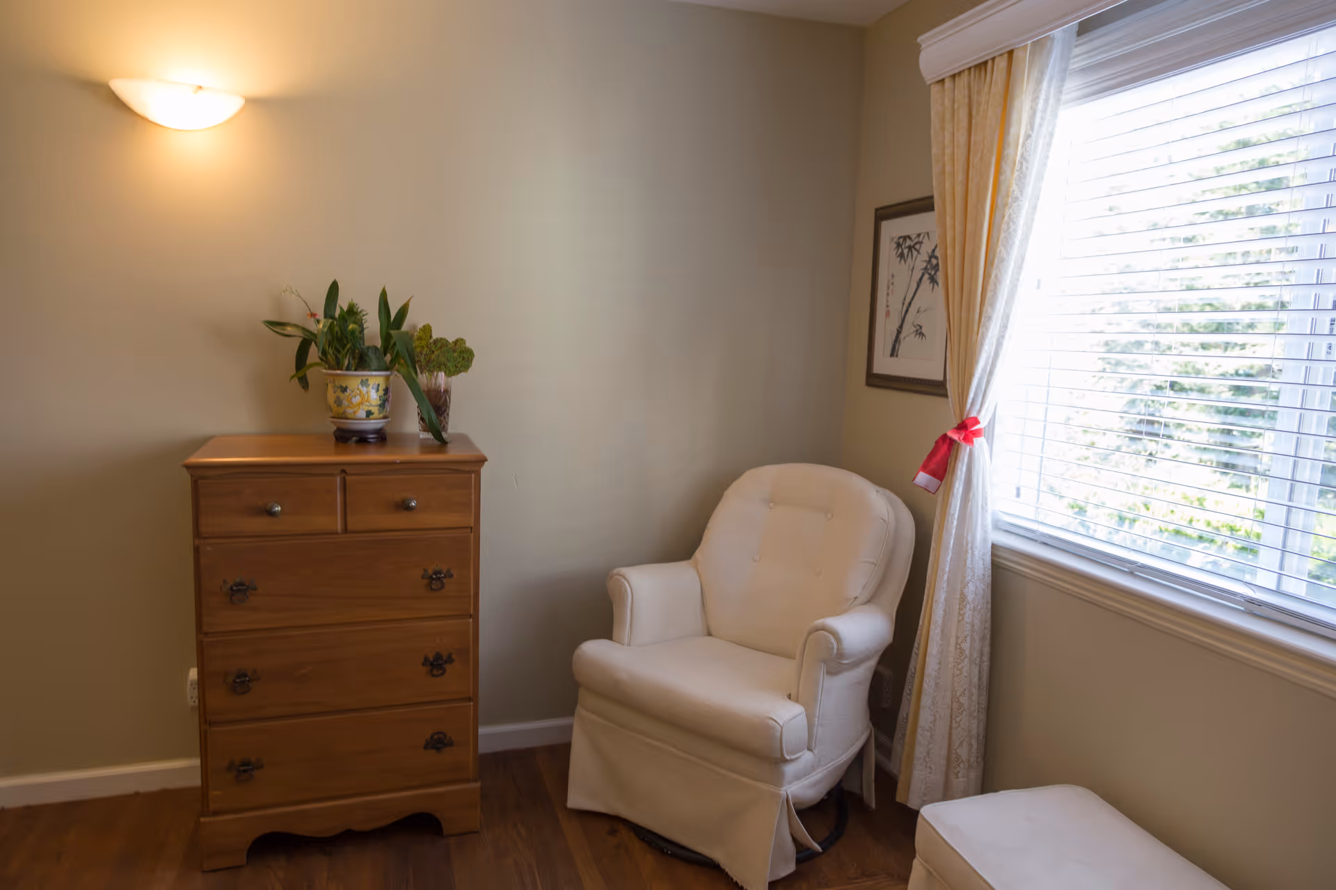 A cozy corner of a room with a white upholstered armchair next to a window with white blinds and cream-colored curtains tied with a red ribbon. To the left of the chair is a wooden chest of drawers with two potted plants on top. The walls are painted beige and there is a framed artwork hanging near the window. The floor is wooden.