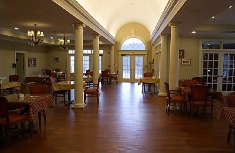 A spacious dining room with wooden floors, multiple tables covered with tablecloths, and chairs arranged around them. The room features large white columns, chandeliers hanging from the ceiling, and a large arched window above double doors at the far end letting in natural light.