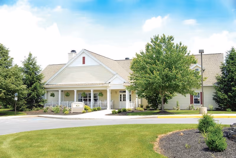 Exterior view of a single-story senior living facility building with a beige facade, white trim, and a covered porch with white railings. The building is surrounded by green trees, shrubs, and a well-maintained lawn under a partly cloudy blue sky.