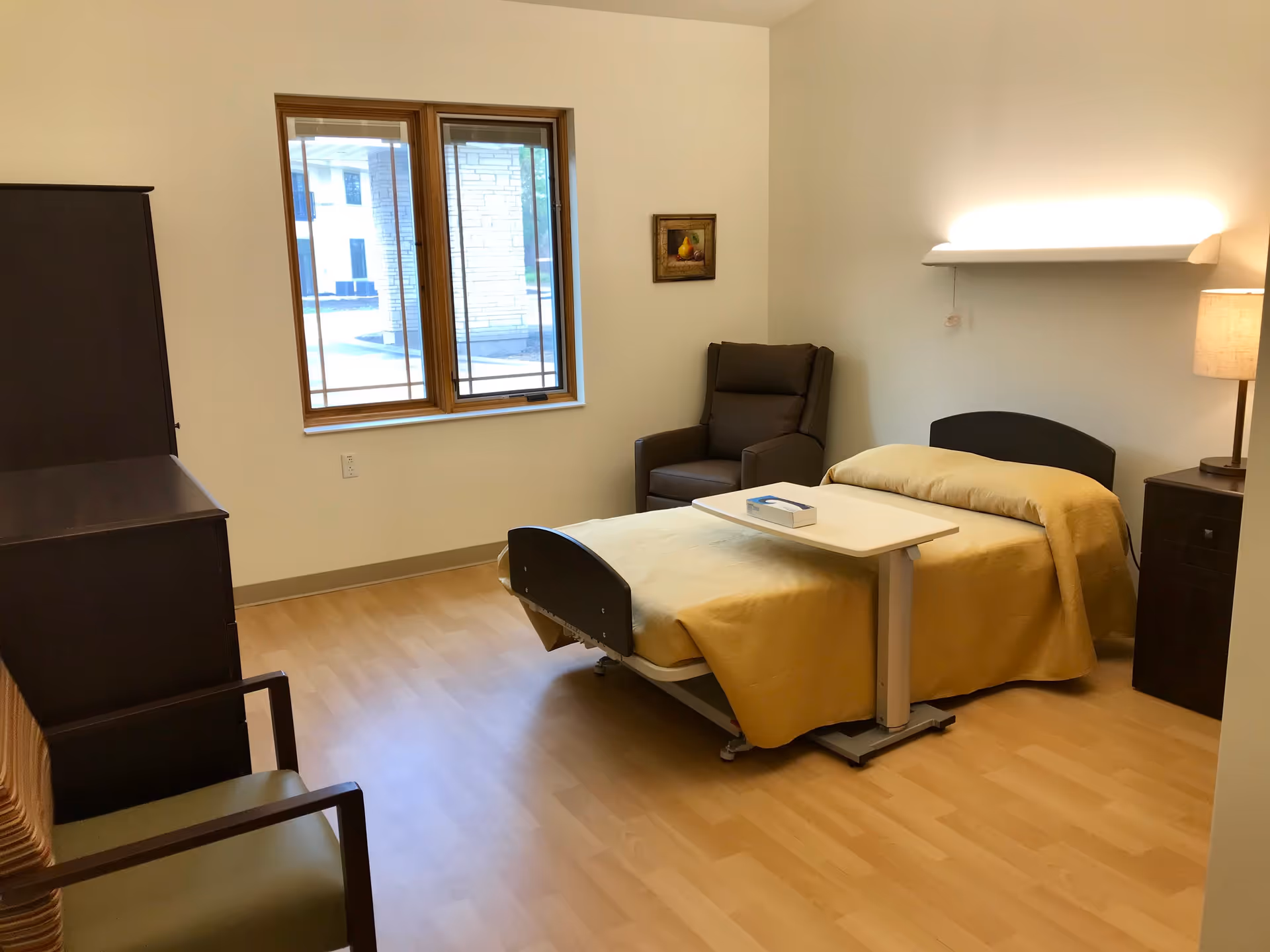 A senior living facility bedroom with a single bed covered in a yellow blanket, a small overbed table with a box of tissues, a dark armchair in the corner, a wooden dresser, a chair with green upholstery, a window with a view outside, a small framed picture on the wall, and a lamp on a nightstand.