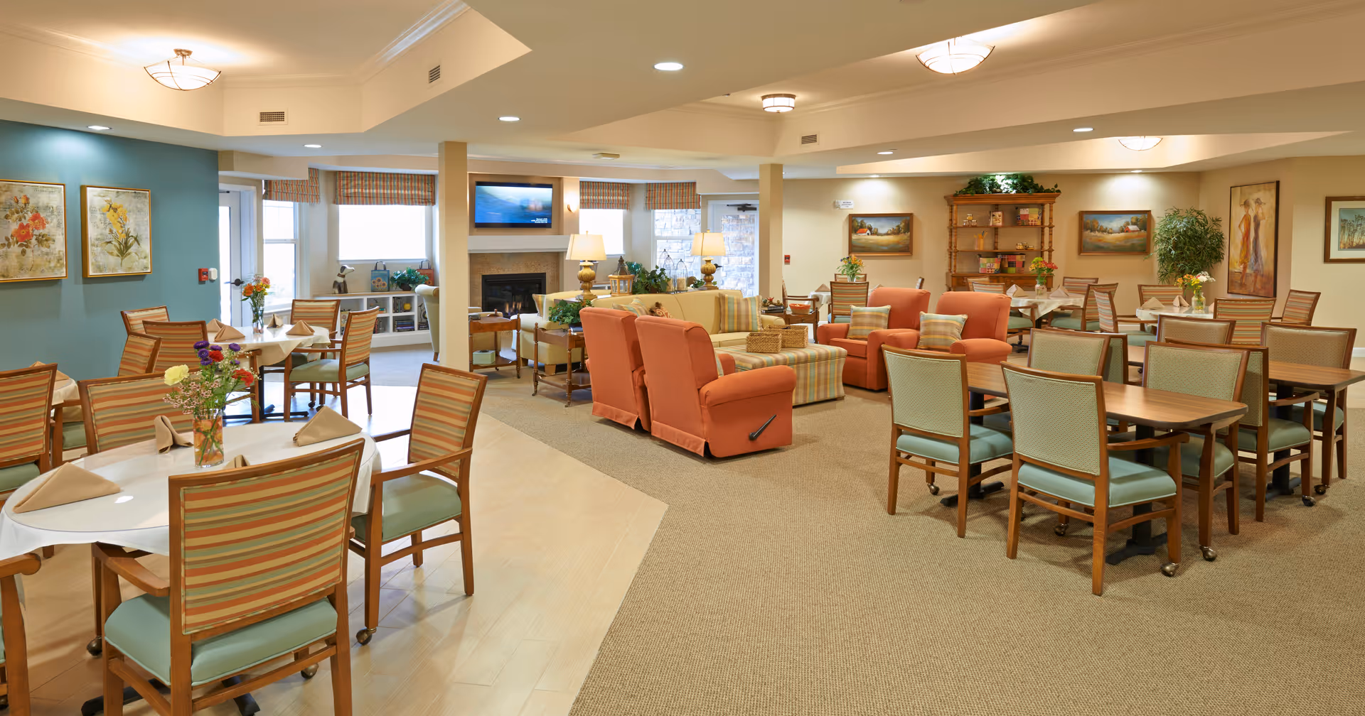 A spacious and well-lit common area in an assisted living facility featuring multiple dining tables with chairs, some set with napkins and flower vases. The room also includes a seating area with orange armchairs and a striped sofa arranged around a fireplace with a TV mounted above it. The walls are decorated with paintings and there are plants placed around the room, creating a warm and inviting atmosphere.