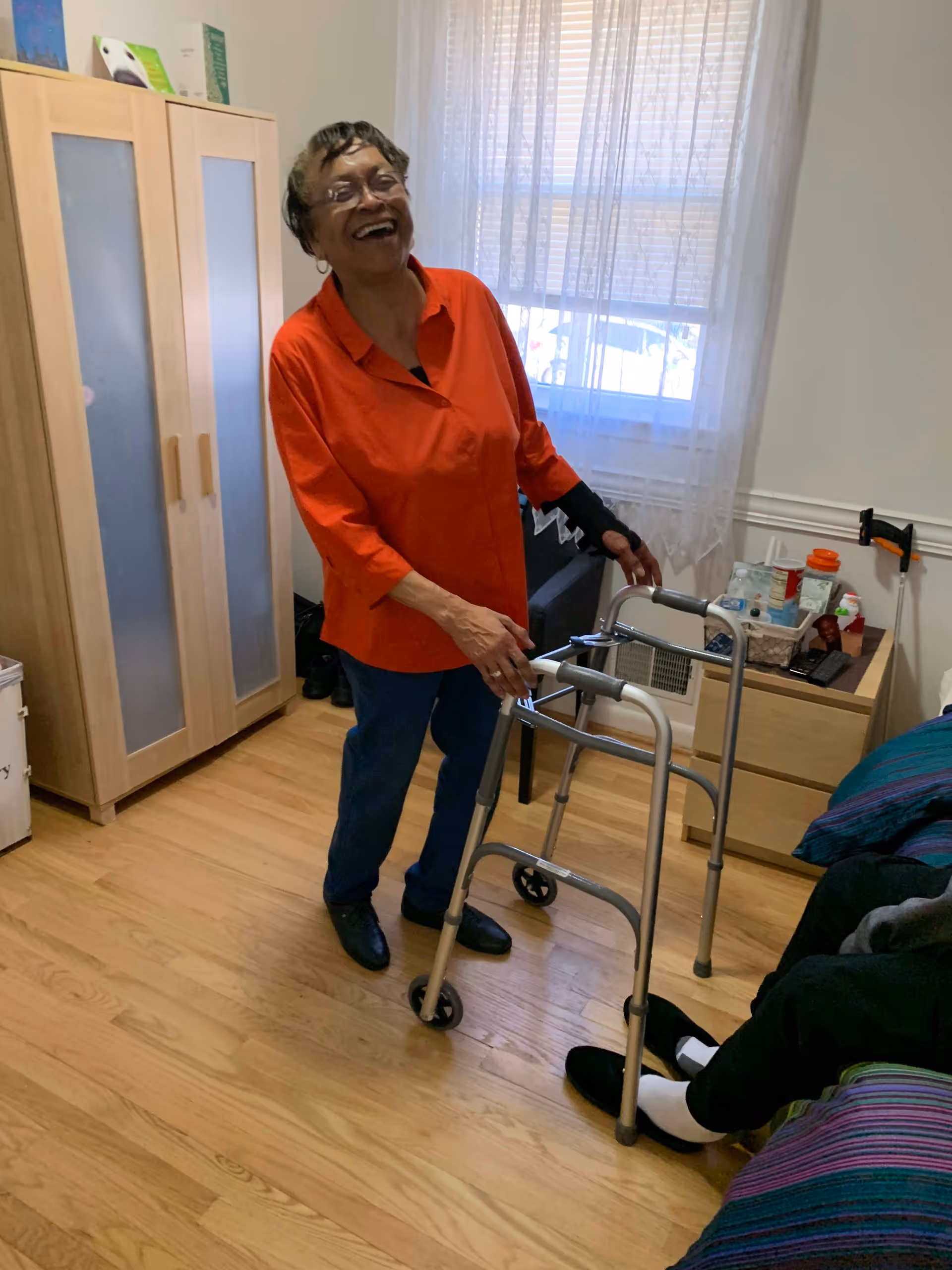 An elderly woman wearing an orange shirt and blue pants is standing and smiling while holding a walker in a room with wooden flooring. There is a wardrobe with frosted glass doors, a window with sheer curtains, and a bedside table with various items including a cane leaning against the wall. A person is sitting on a bed partially visible on the right side of the image.