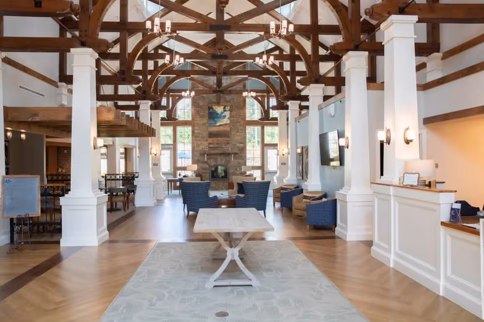 Spacious senior living lobby with exposed wooden beams, a central table, seating areas, fireplace and a reception desk.