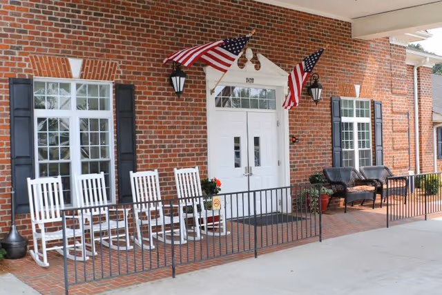 Front entrance of a brick building with two American flags mounted above a white double door. There are four white rocking chairs and two black wicker chairs with cushions on a brick patio, separated from the driveway by a black metal railing.