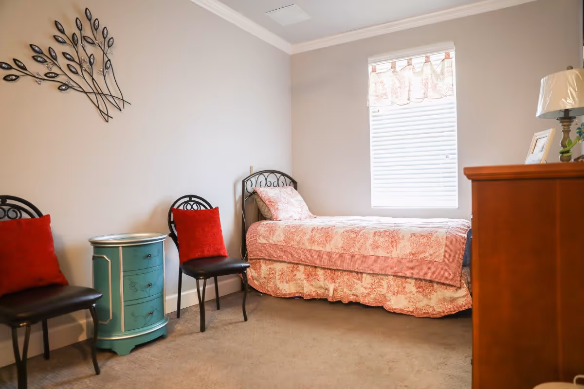 A small bedroom with a single bed covered in a pink and white patterned quilt, two black chairs with red cushions, a small turquoise nightstand between the chairs, a wooden dresser with a lamp and picture frame, and a window with white blinds and a pink valance.