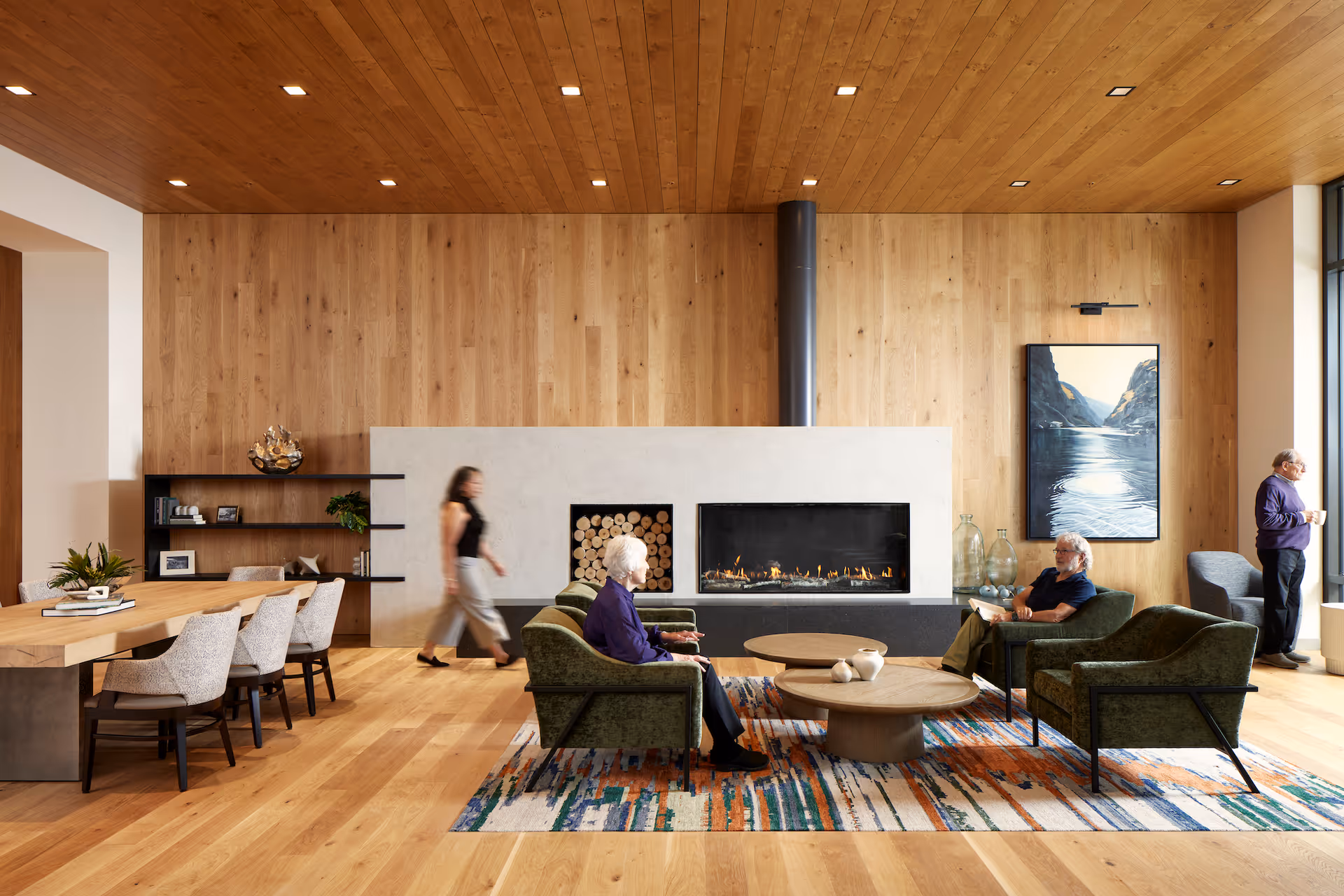 A modern senior living facility common area with wooden floors and ceiling, a large fireplace with stacked firewood, green armchairs arranged around two round wooden coffee tables on a colorful rug. Three seniors are seated and conversing, while a woman walks past a dining table with chairs on the left. Large windows on the right let in natural light, and a landscape painting hangs on the wooden wall above the fireplace.