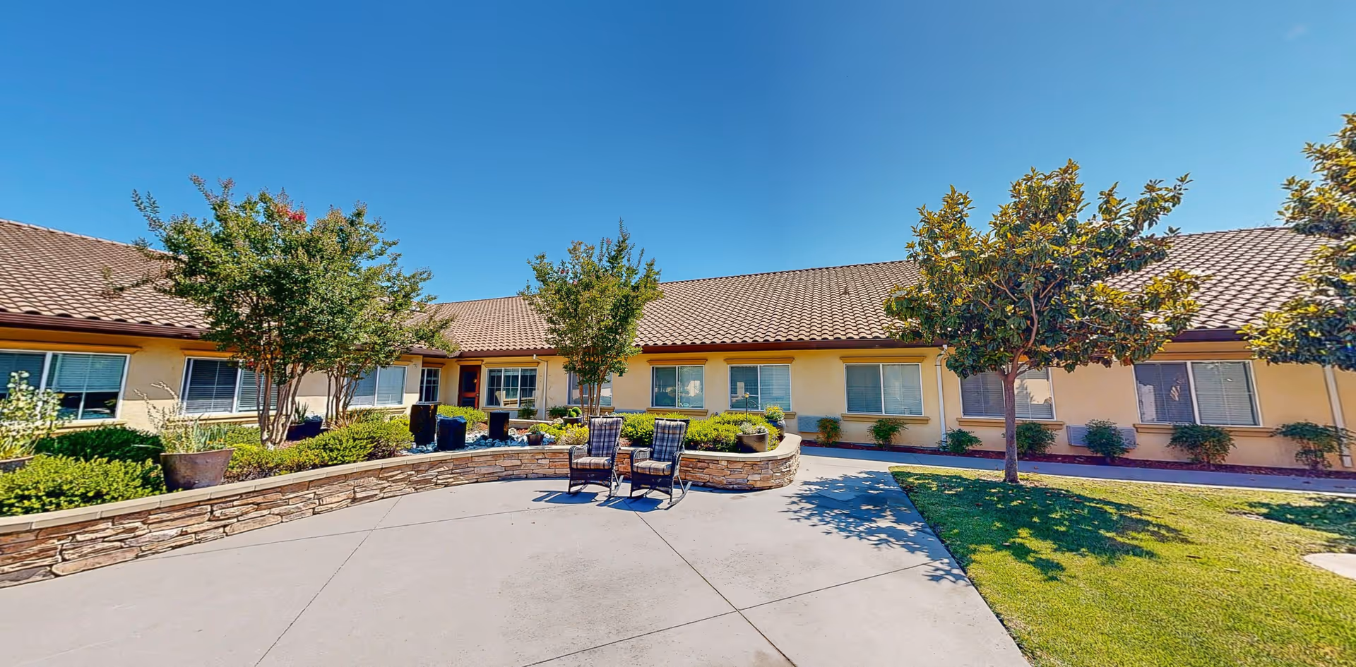 Outdoor courtyard area of Cogir of Folsom Memory Care with a paved patio, two chairs, trees, shrubs, and a single-story building with multiple windows under a clear blue sky.