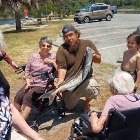 A group of people outdoors near a parking lot, including elderly individuals sitting in wheelchairs and a man kneeling while holding a large fish. There are trees, picnic tables, and a parked car in the background.