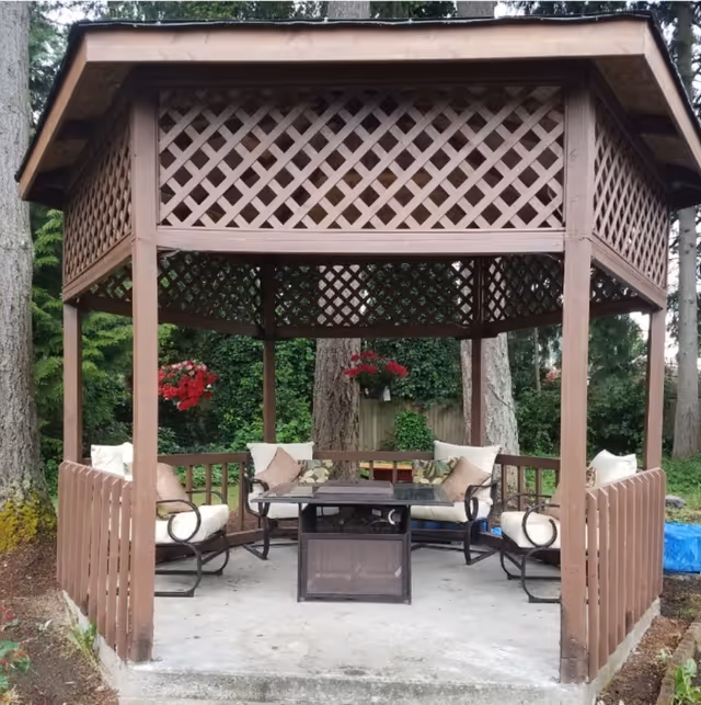 Octagonal wooden gazebo with cushioned patio chairs, a central table, and hanging flower baskets in a wooded yard.