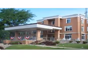 Exterior view of a three-story brick building with a covered entrance and a ramp, decorated with red, white, and blue bunting. There is a grassy area and some landscaping in front of the building.