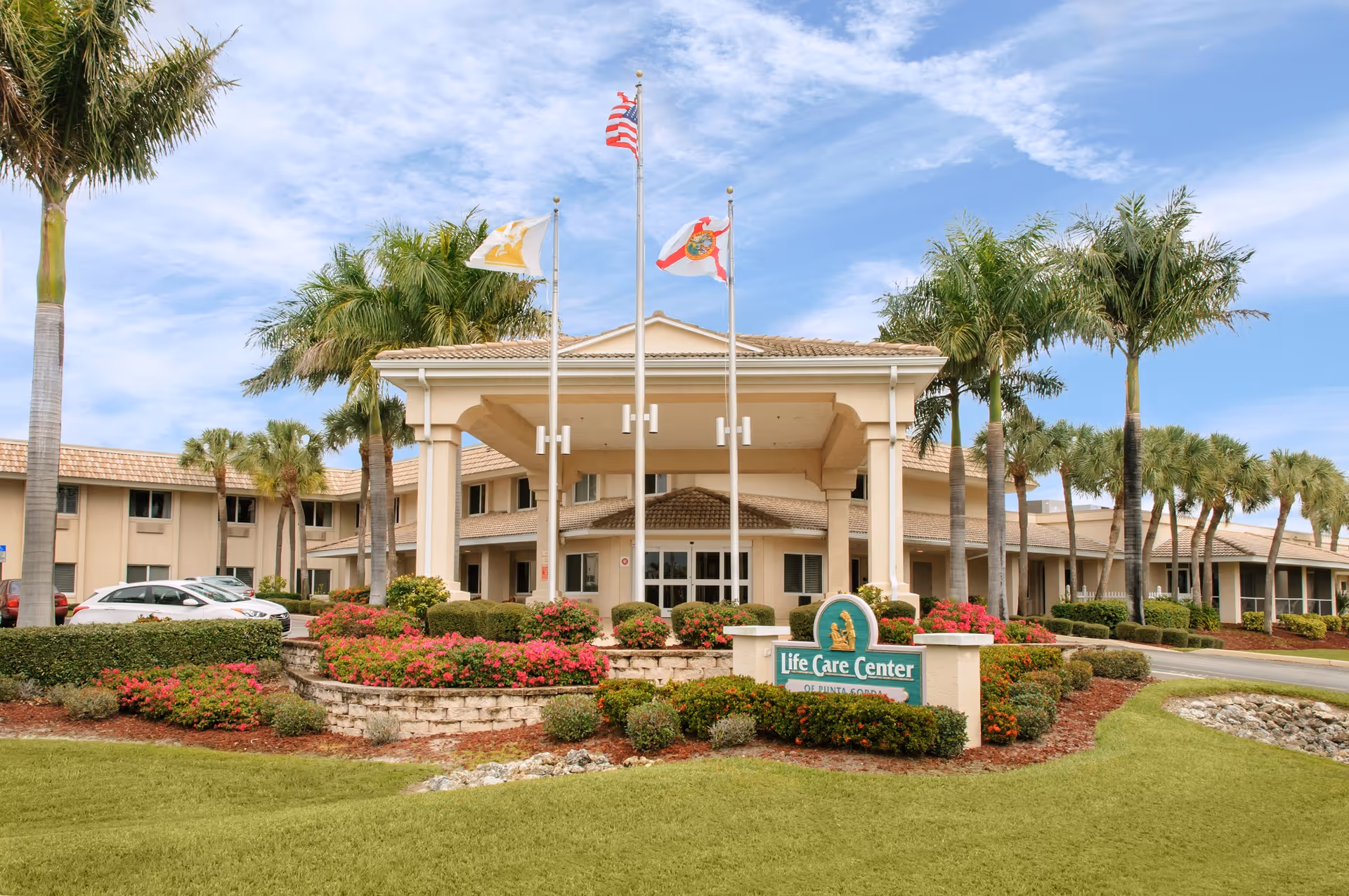 Front exterior view of Life Care Center of Punta Gorda building with a covered entrance, three flagpoles flying the American flag, Florida state flag, and another flag. The building is surrounded by palm trees, well-maintained landscaping with green grass, bushes, and colorful flowers under a partly cloudy sky.