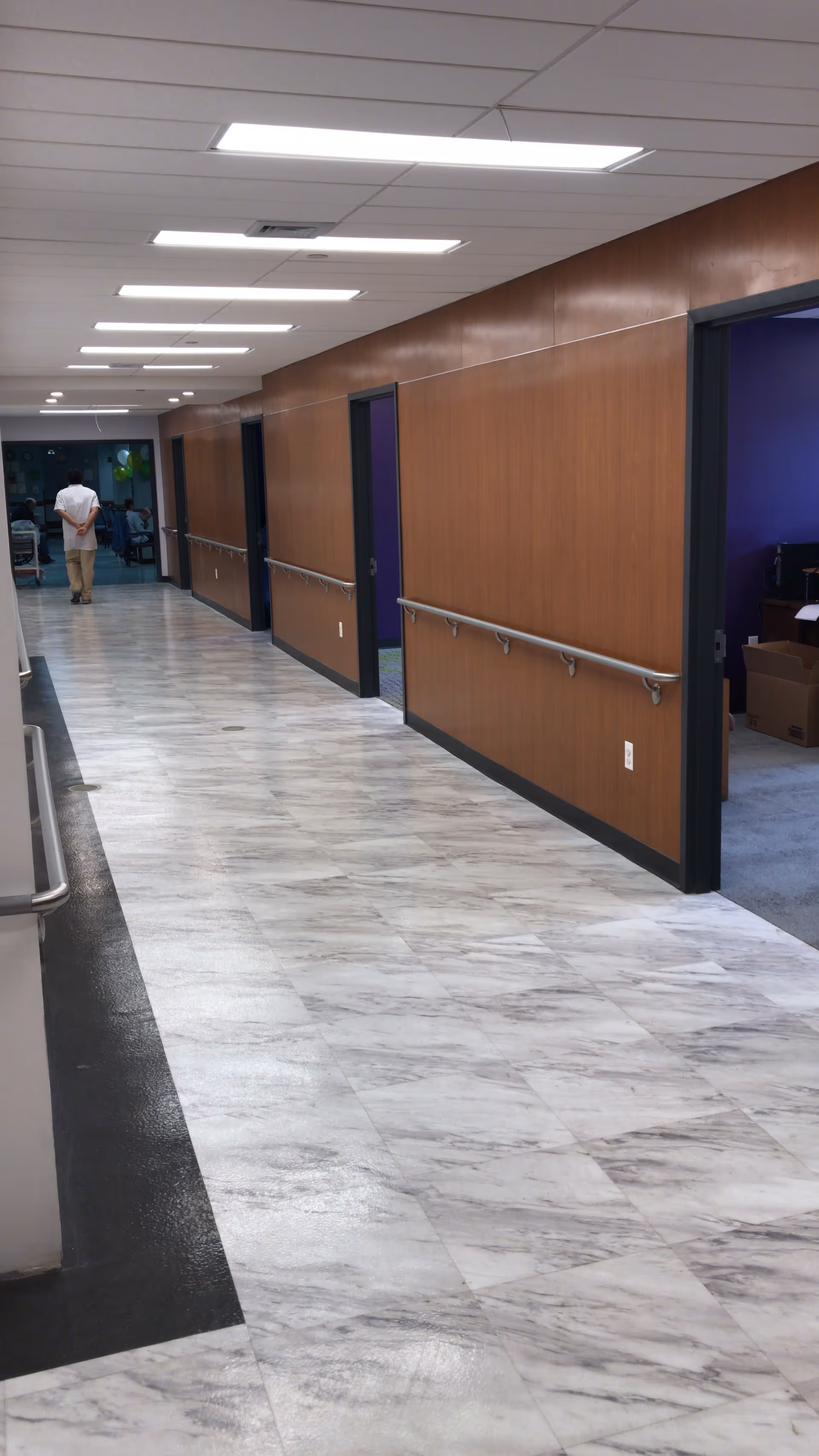 A long, clean hallway in a senior living facility with marble-patterned flooring, wooden panel walls, handrails on both sides, and several open doorways. A person is walking away at the end of the hallway.