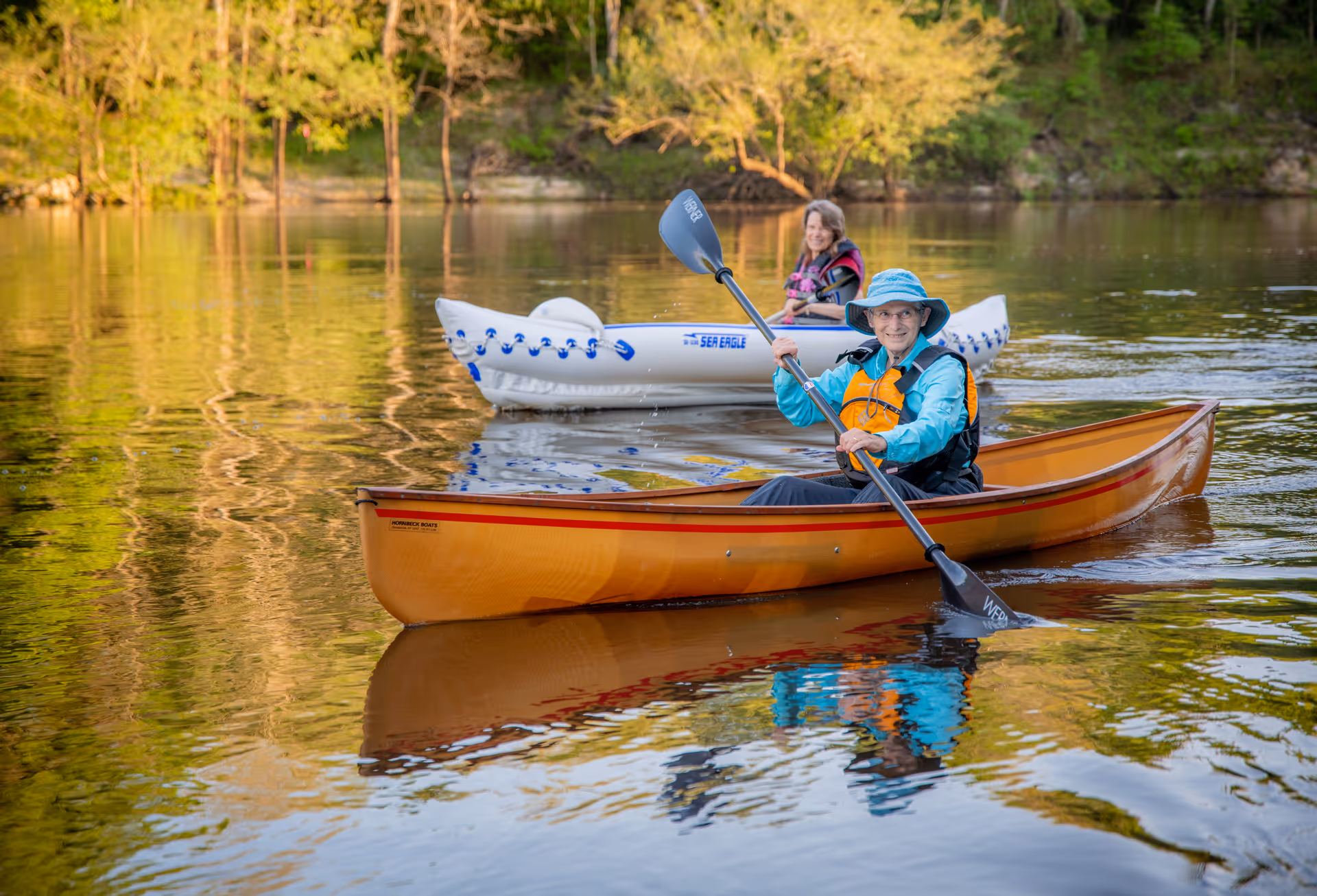 Two people wearing life jackets paddle a canoe and an inflatable kayak on a calm, tree-lined lake.