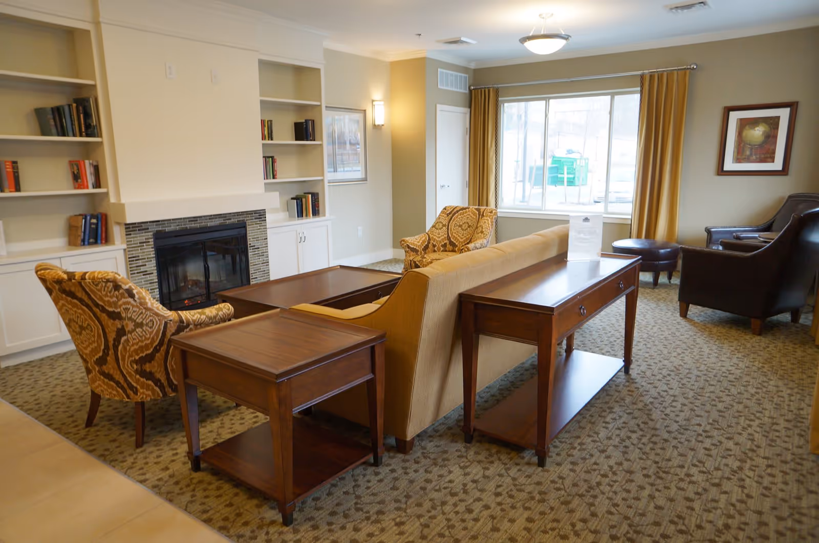 A cozy living room area with a beige sofa, patterned armchairs, dark leather chairs, wooden side tables, a fireplace with built-in shelves filled with books, a large window with yellow curtains, and framed artwork on the walls.