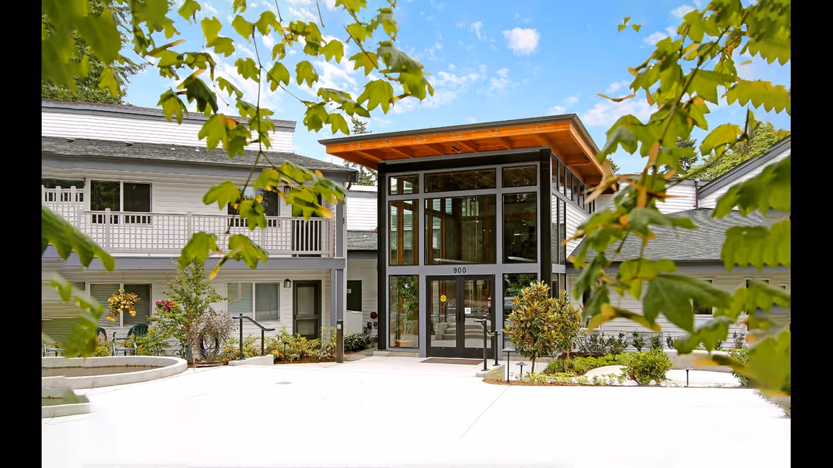 Front entrance of a senior living building featuring a two-story glass atrium, surrounding landscaping, and tree branches in the foreground.