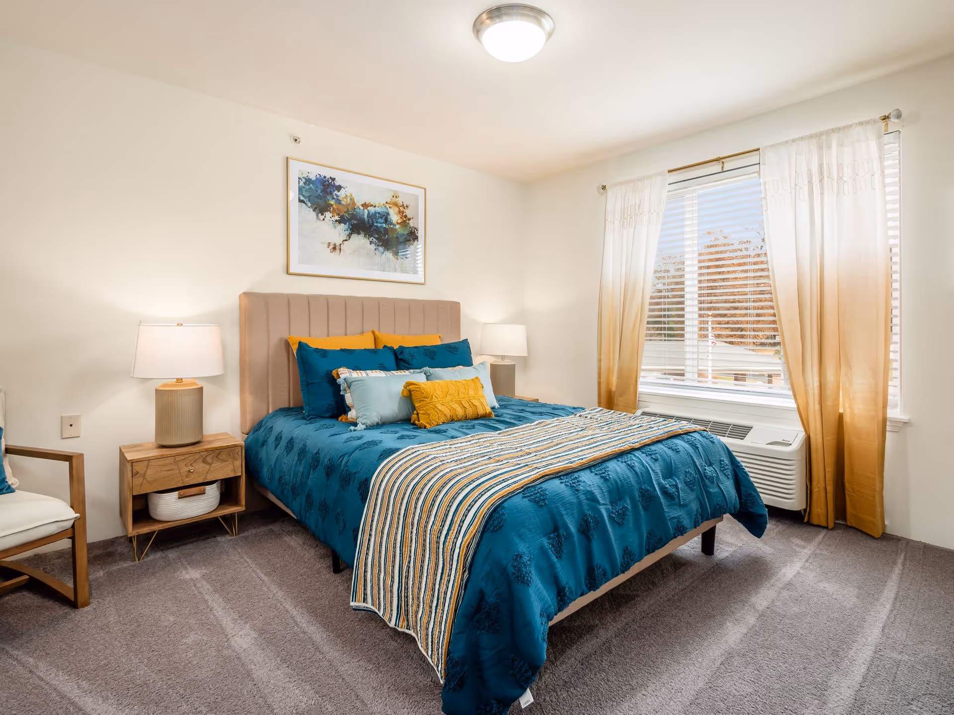 A neatly made bed with teal bedding and multiple pillows in a bedroom. There are two wooden nightstands with lamps on either side of the bed. A large window with sheer beige curtains lets in natural light. A framed abstract painting hangs above the bed. The room has beige walls and carpeted flooring.