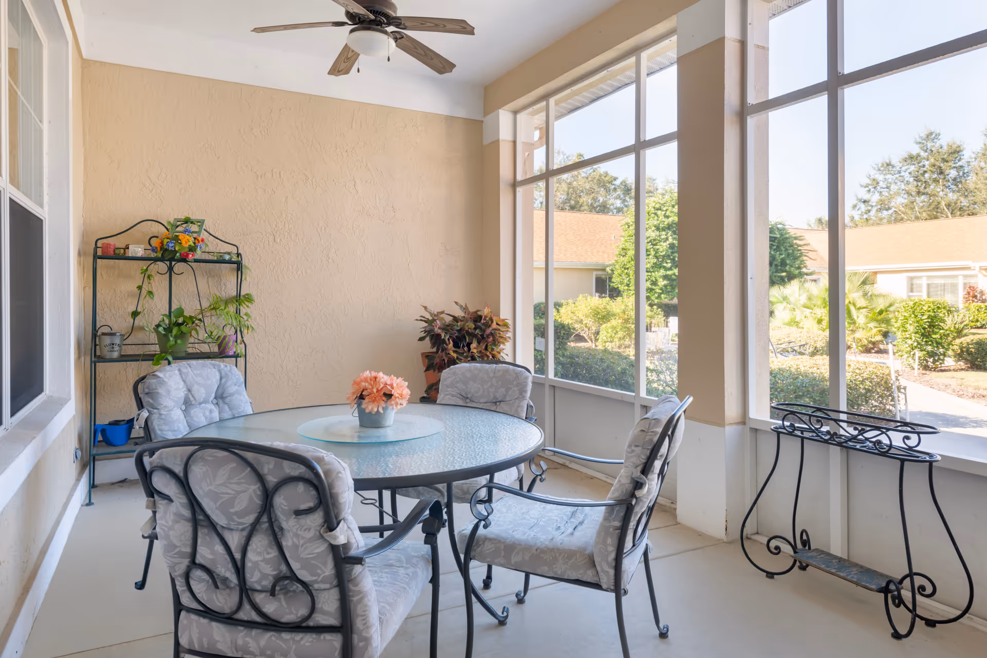 A screened-in patio area with a round glass table surrounded by four cushioned metal chairs. There is a ceiling fan above, a small metal shelf with potted plants against the wall, and a decorative metal plant stand near the screened windows. Outside the screened area, greenery and neighboring buildings are visible.